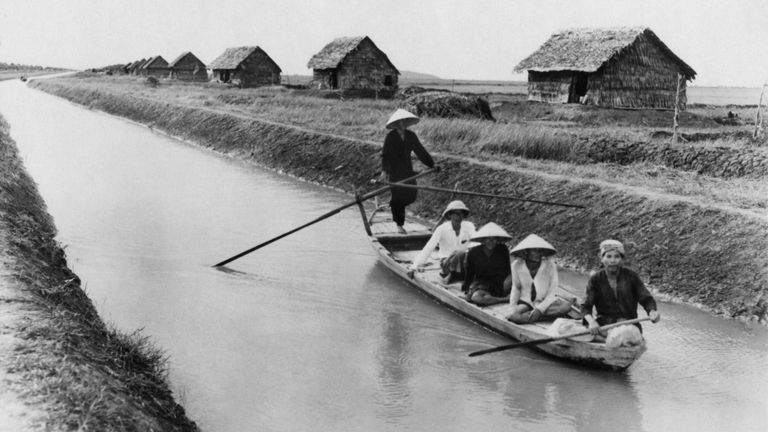 Vietnamese men in a sampan on a canal near straw dwellings that house refugees from North Vietnam in 1956. | Source: Shutterstock